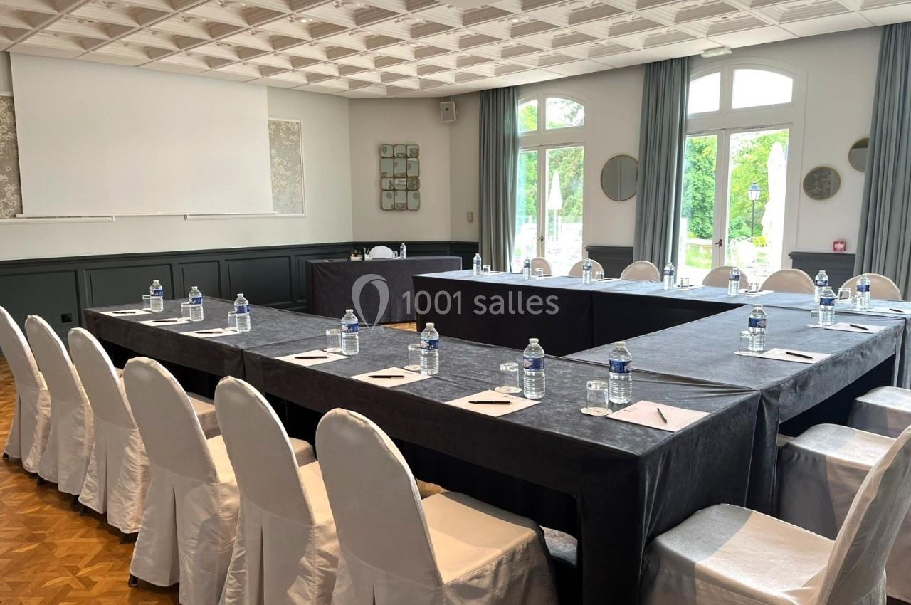 Salle de réunion lumineuse avec tables disposées en U, chaises blanches, bouteilles d'eau et blocs-notes.