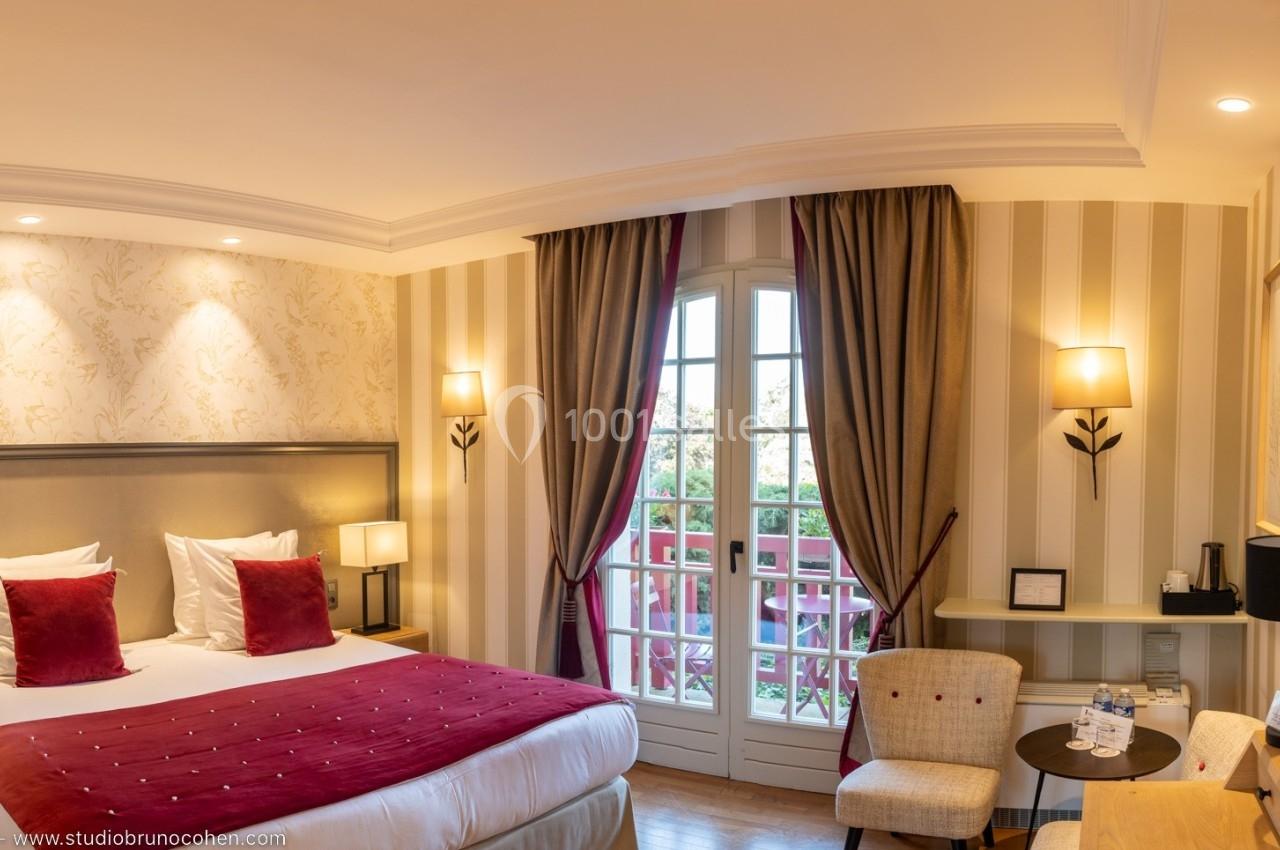 Chambre d'hôtel lumineuse avec lit double, coussins rouges, bureau, télévision et fenêtre donnant sur un balcon fleuri.