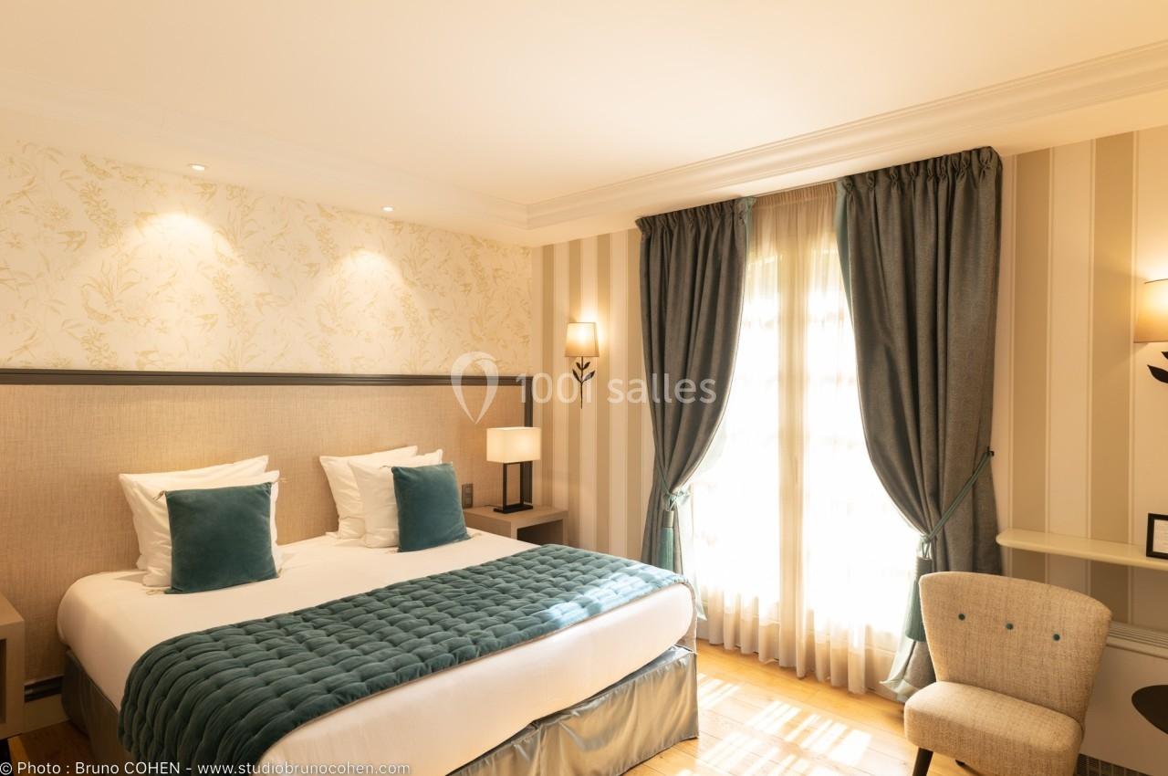 Chambre lumineuse avec lit double, coussins bleus, rideaux assortis, fauteuil et bureau près d'une fenêtre.