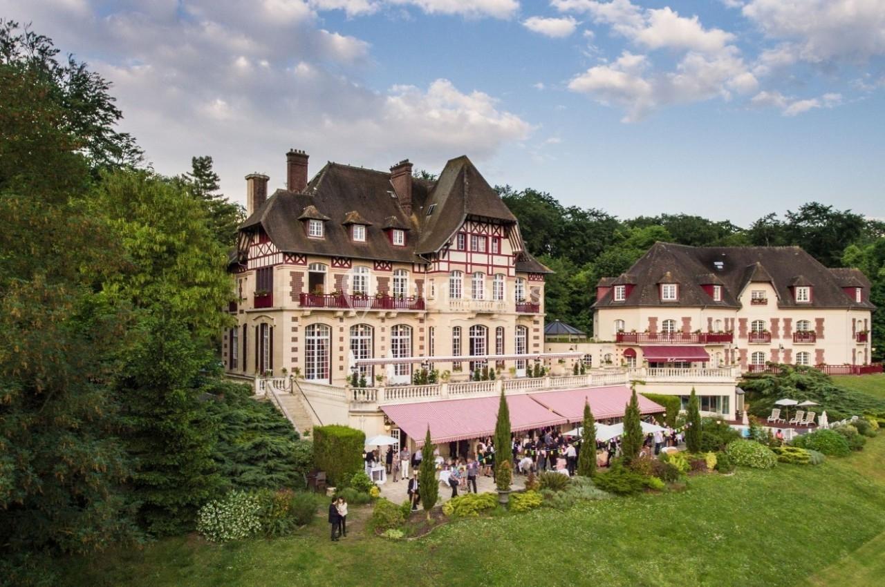 Vue aérienne d'un manoir entouré de verdure avec une terrasse animée et un ciel partiellement nuageux.