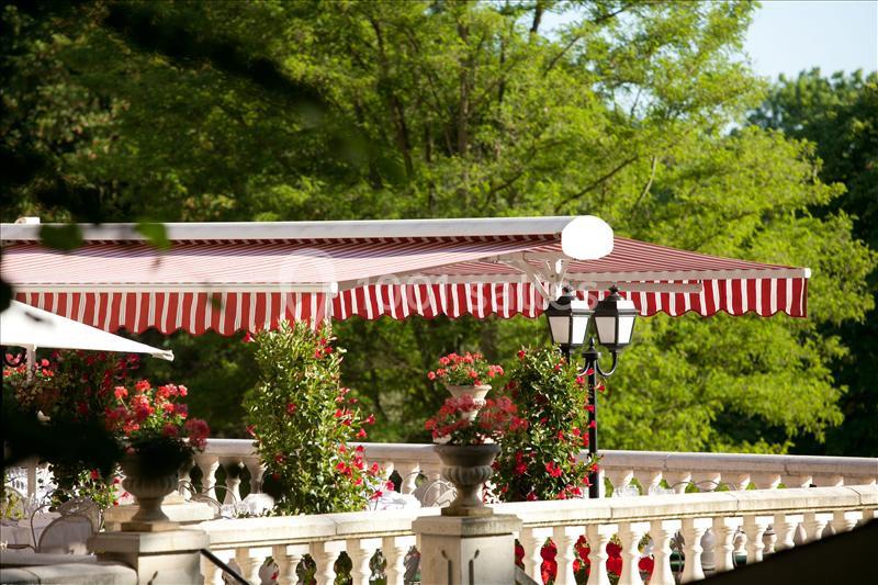 Terrasse avec balustrade, stores rayés rouges et blancs, entourée de plantes fleuries, devant un fond arboré.
