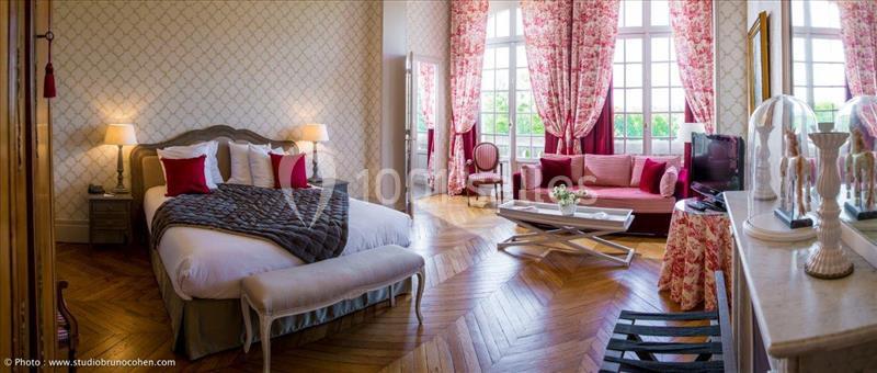 Chambre élégante avec lit double, canapé, grandes fenêtres, parquet en bois et décoration classique aux tons rouges et…