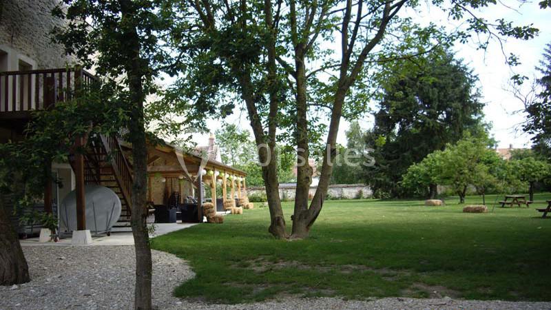 Jardin verdoyant avec pelouse, arbres, tables de pique-nique et terrasse en bois attenante à un bâtiment en pierre.
