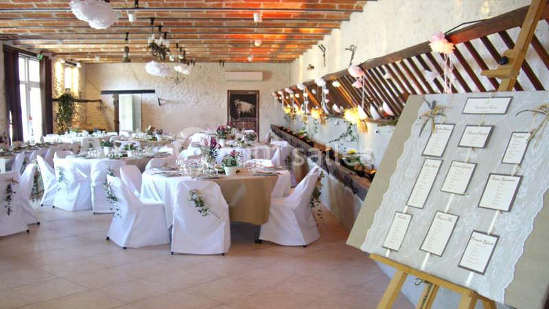 Salle de réception décorée pour un mariage avec tables dressées, chaises blanches et plan de table sur un chevalet.