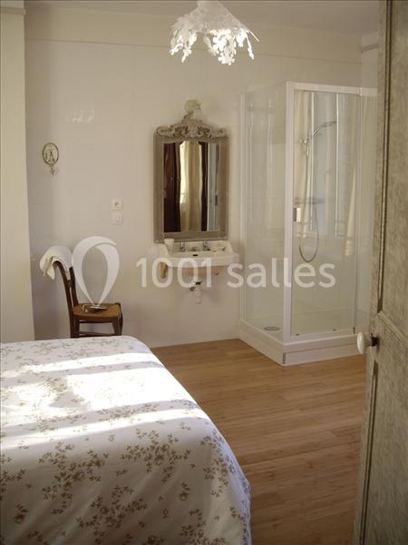 Chambre lumineuse avec lit à motifs floraux, chaise en bois, lavabo avec miroir et cabine de douche vitrée.