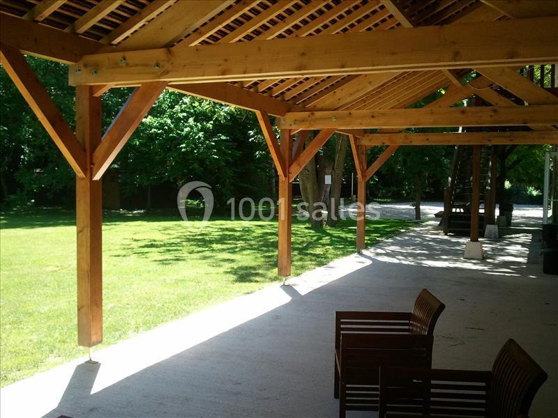 Structure en bois ouverte avec toit incliné, donnant sur un espace vert arboré et des bancs en bois au premier plan.