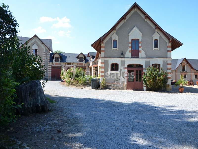 Grande maison en pierre avec des volets rouges, entourée de gravier, de verdure et d'arbustes fleuris.
