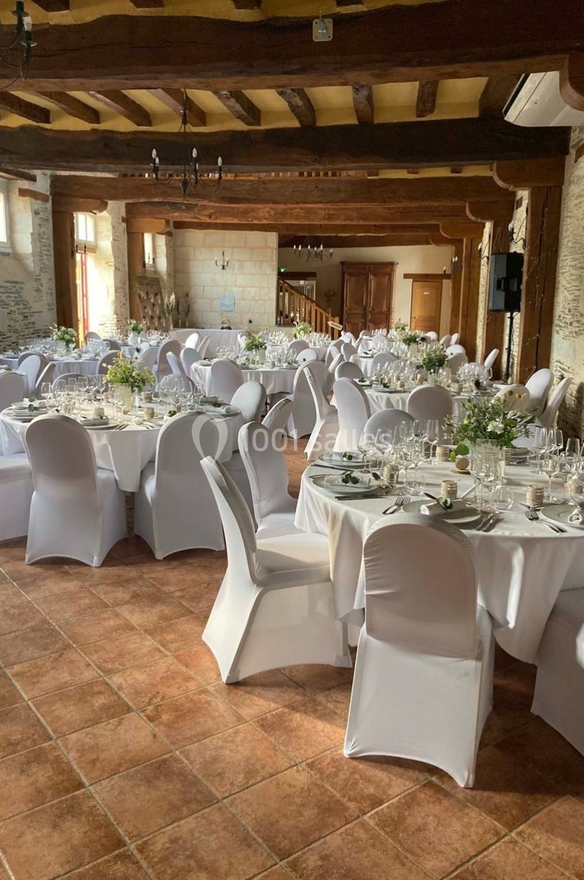 Salle de réception avec tables rondes dressées, nappes blanches, chaises houssées et décorations florales.