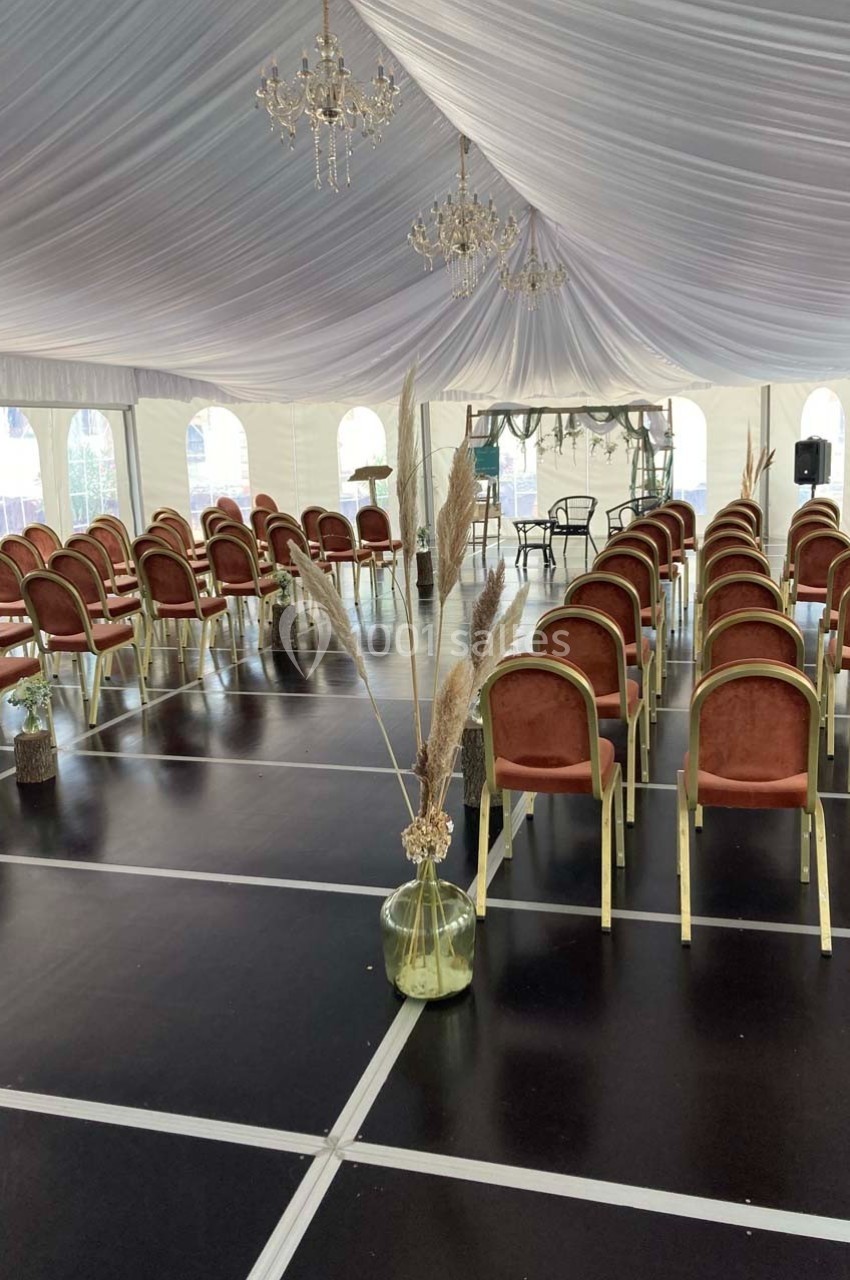 Chaises alignées sous une tente blanche avec un sol noir, décorée de pampas dans des vases et de lustres suspendus.