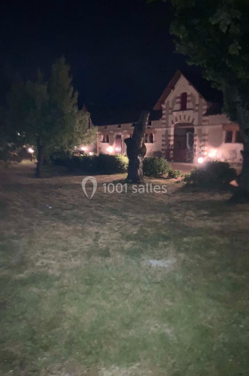 Maison ancienne éclairée la nuit, entourée d'arbres et d'un jardin avec pelouse.