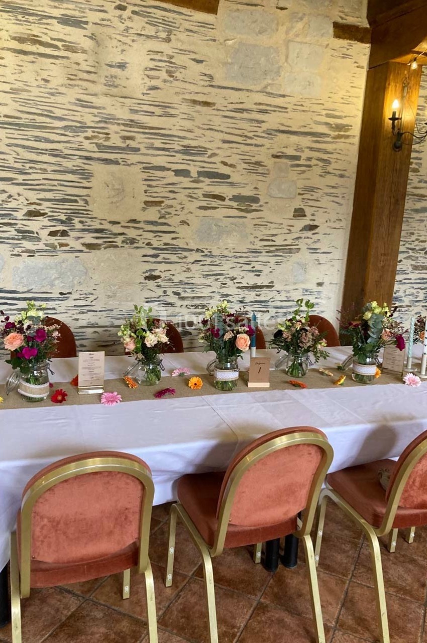 Table décorée pour un événement, avec nappes blanches, chaises rouges et vases de fleurs colorées devant un mur en pierre.