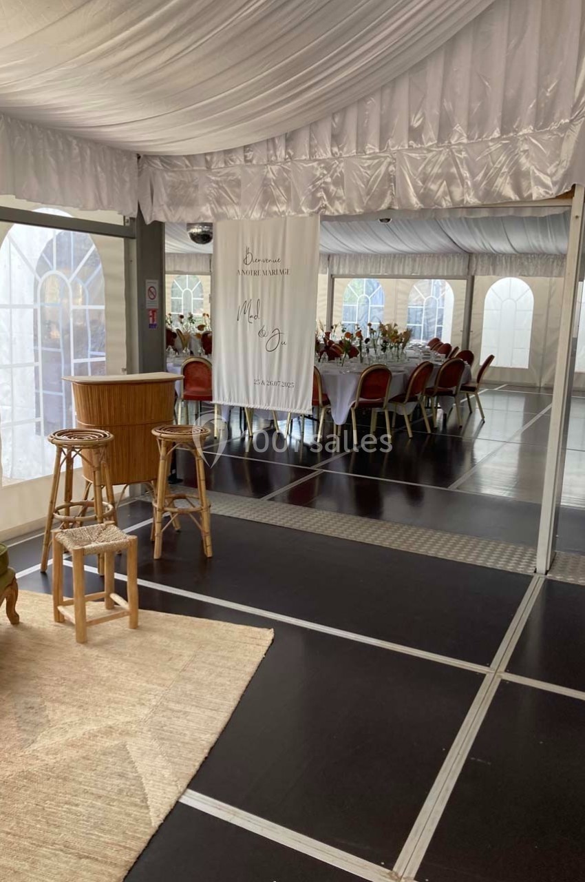 Salle de réception décorée avec des tables dressées, des chaises rouges et un panneau de bienvenue suspendu.