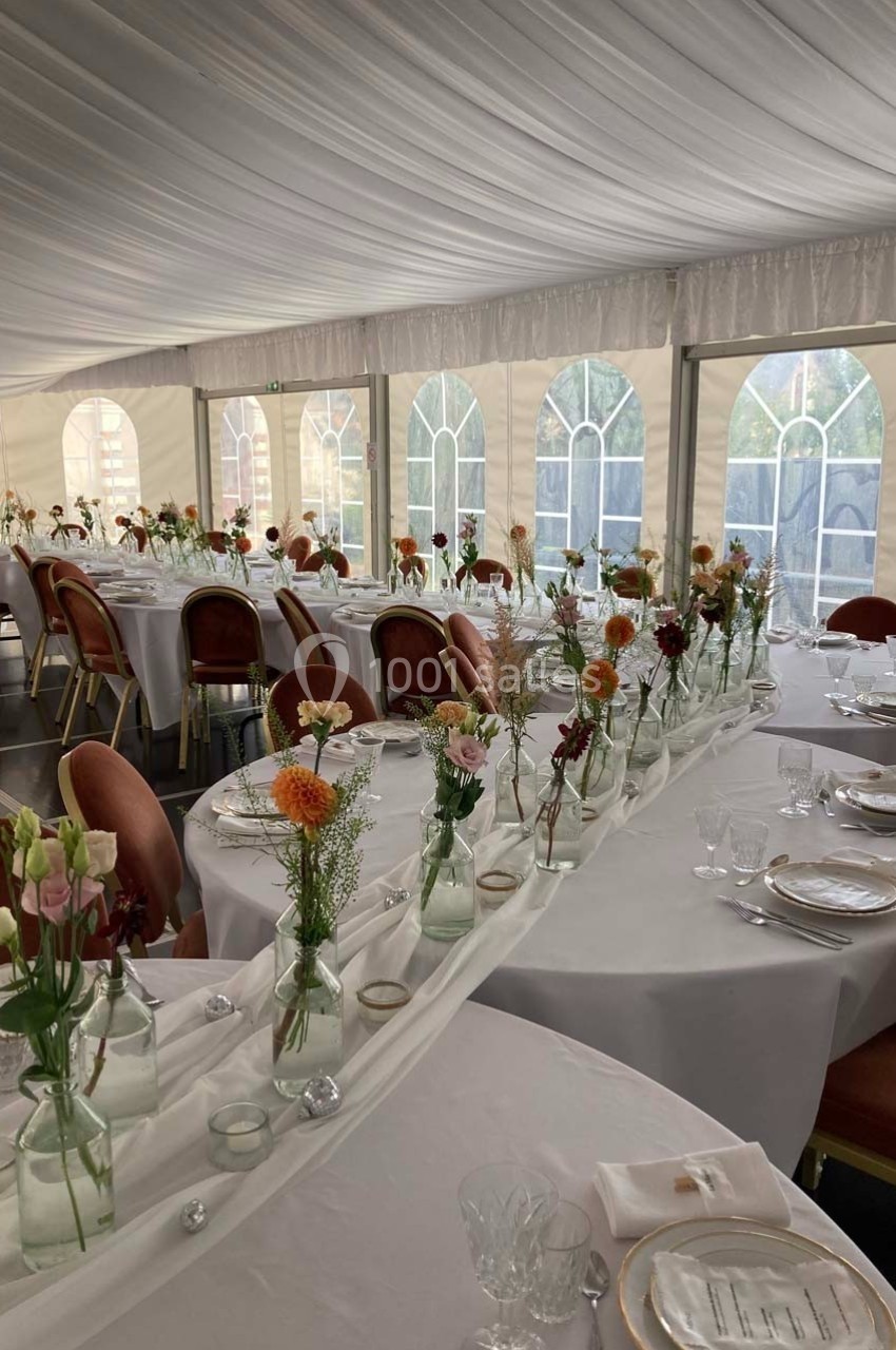 Salle de réception décorée avec des tables rondes, nappes blanches et bouquets de fleurs dans des vases en verre.