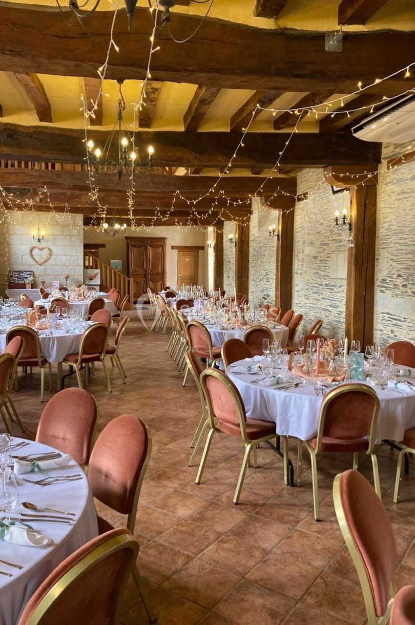 Salle de réception décorée avec des tables rondes dressées, chaises rouges, poutres apparentes et guirlandes lumineuses.