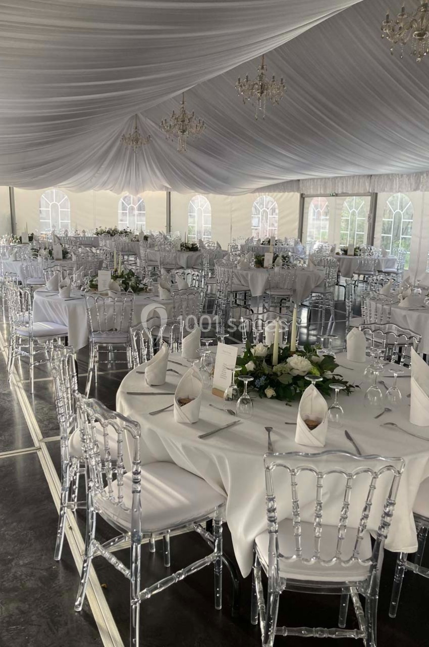 Salle de réception sous chapiteau avec tables rondes dressées, nappes blanches et chaises transparentes.