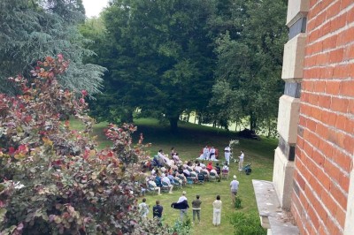 Groupe de personnes assises en cercle dans un jardin verdoyant, vu depuis une fenêtre d'un bâtiment en briques.