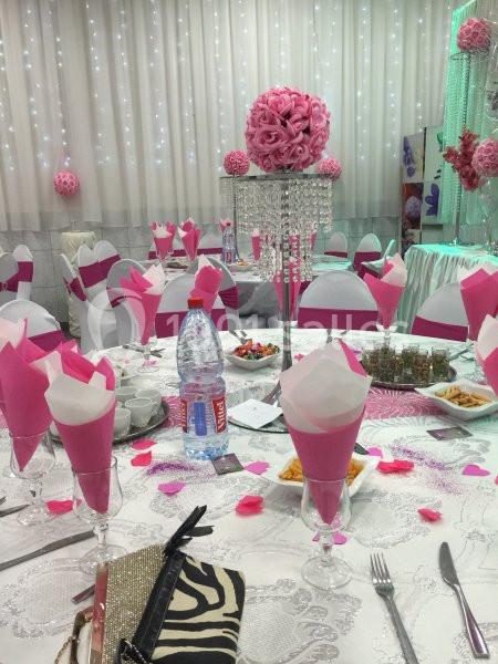 Salle de réception décorée en rose et blanc avec tables dressées, nappes ornées et centre de table floral.