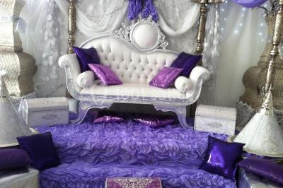 Deux femmes en robes traditionnelles violettes ornées de bijoux, posant devant un fond clair avec un fauteuil argenté.