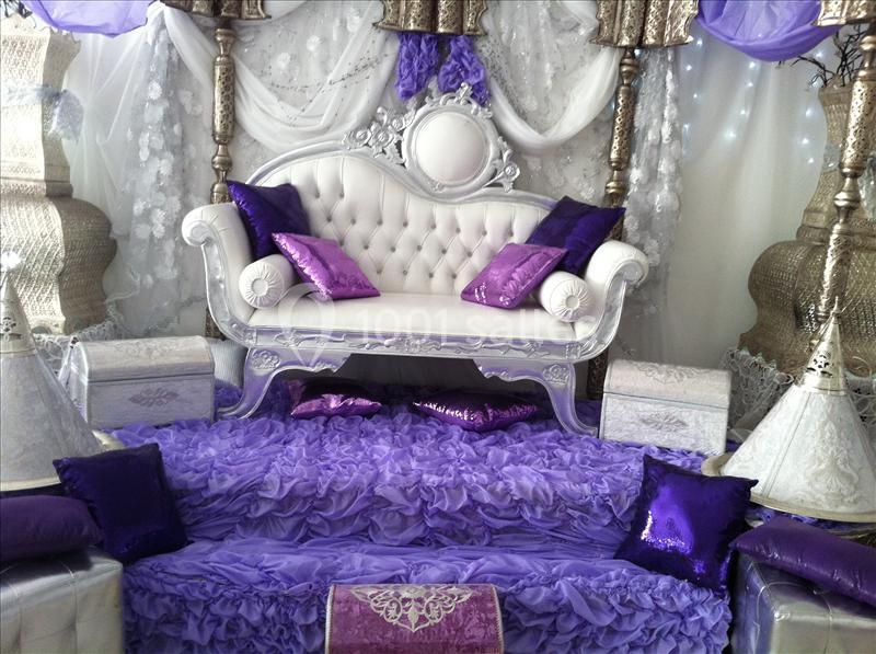 Canapé blanc capitonné entouré de coussins violets et décorations argentées dans un cadre festif et élégant.