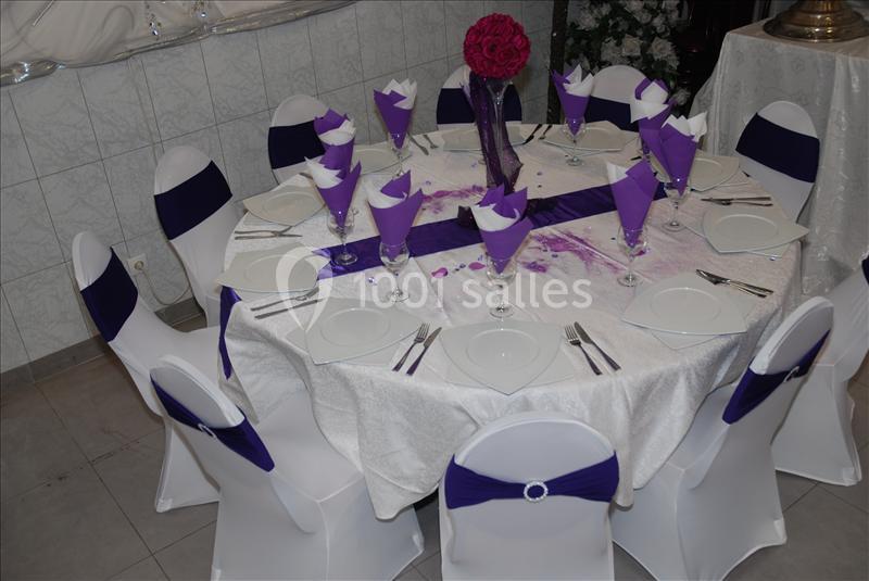 Table ronde dressée pour un événement, avec nappes blanches, serviettes violettes pliées et décoration florale centrale.