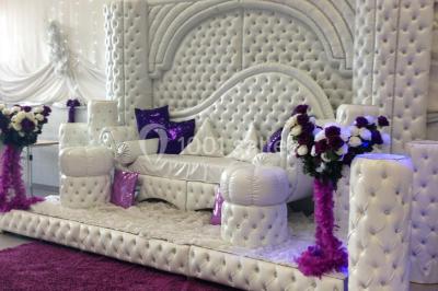 Deux femmes en robes traditionnelles violettes ornées de bijoux, posant devant un fond clair avec un fauteuil argenté.