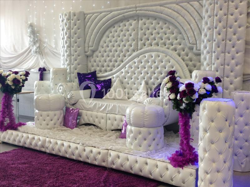 Canapé blanc capitonné avec coussins violets, entouré de colonnes décorées de fleurs et de tissus violets.