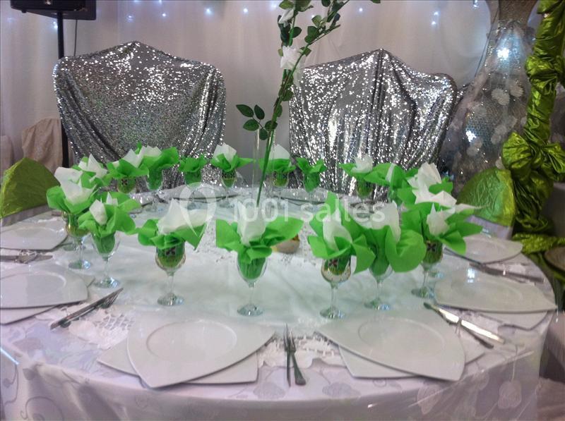 Table ronde décorée pour un événement, avec nappes blanches, serviettes vertes pliées et chaises couvertes de tissu argenté.