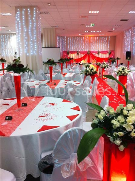 Salle de réception décorée en rouge et blanc avec tables rondes, chaises couvertes et arrangements floraux.