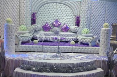 Deux femmes en robes traditionnelles violettes ornées de bijoux, posant devant un fond clair avec un fauteuil argenté.