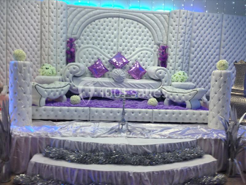 Décor de scène avec canapé capitonné blanc, coussins violets, fleurs décoratives et éclairage lumineux en arrière-plan.