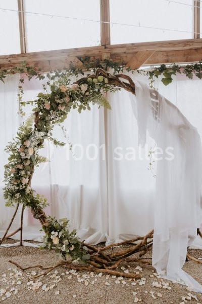 Arche circulaire en bois décorée de fleurs blanches et feuillage, avec un voile blanc, devant des rideaux translucides.