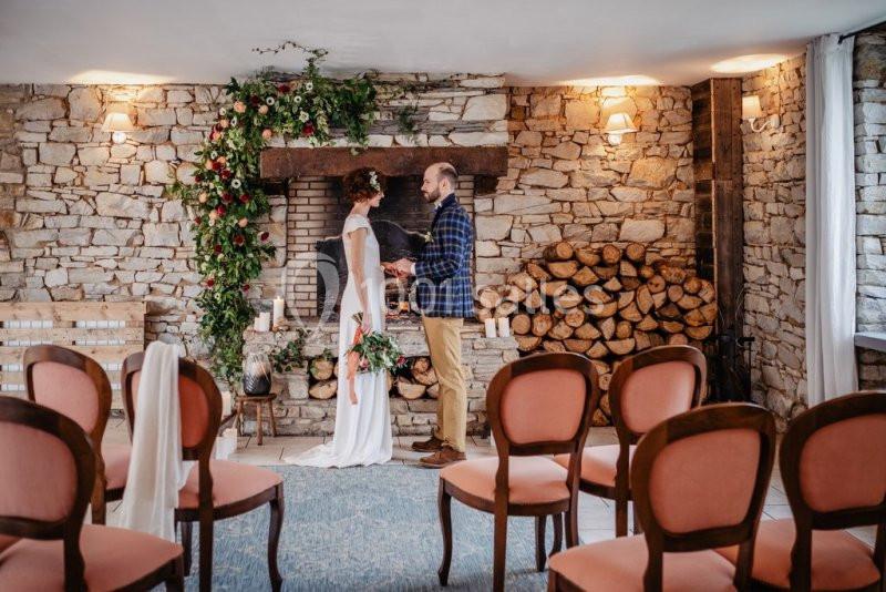 Un couple échange devant une cheminée décorée de fleurs dans une salle en pierre avec des chaises alignées.