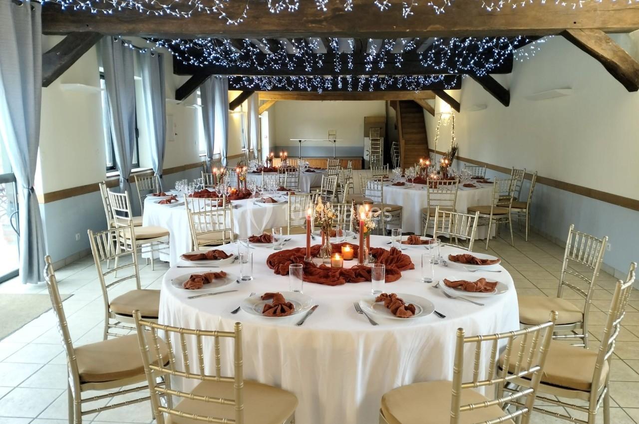 Salle de réception décorée avec des tables rondes, nappes blanches, serviettes rouges et guirlandes lumineuses au plafond.