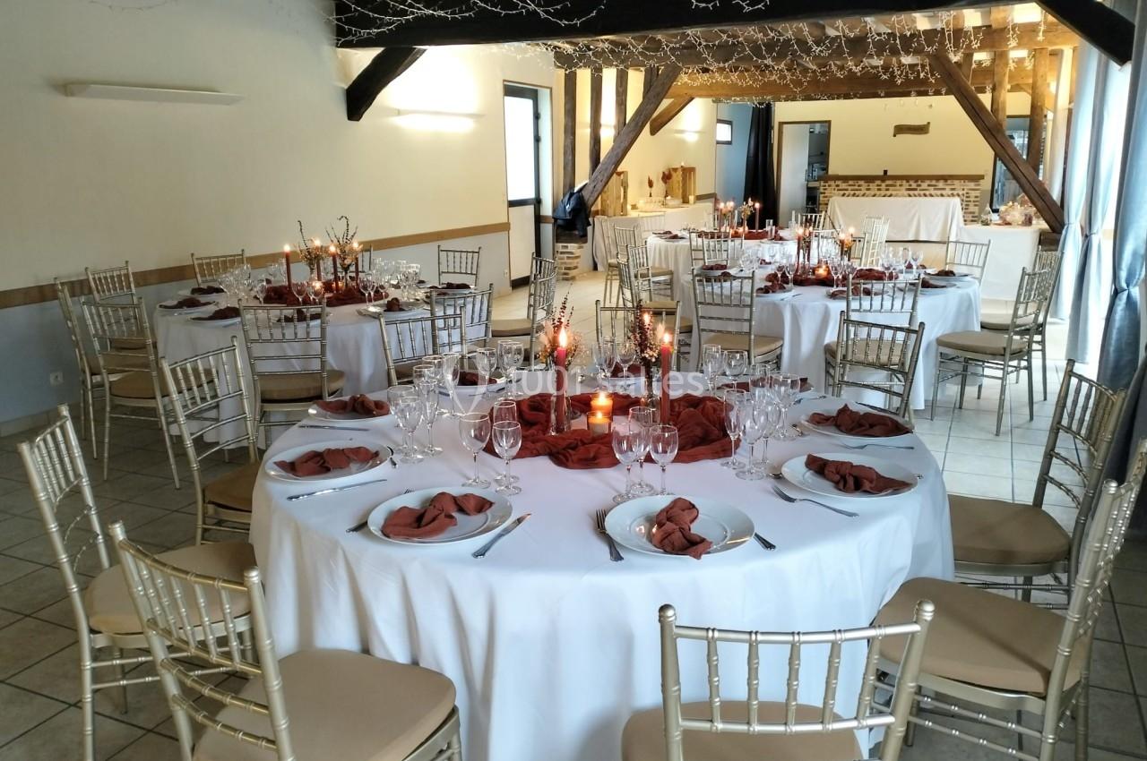 Salle de réception décorée avec des tables rondes, nappes blanches, serviettes rouges et chandeliers allumés.