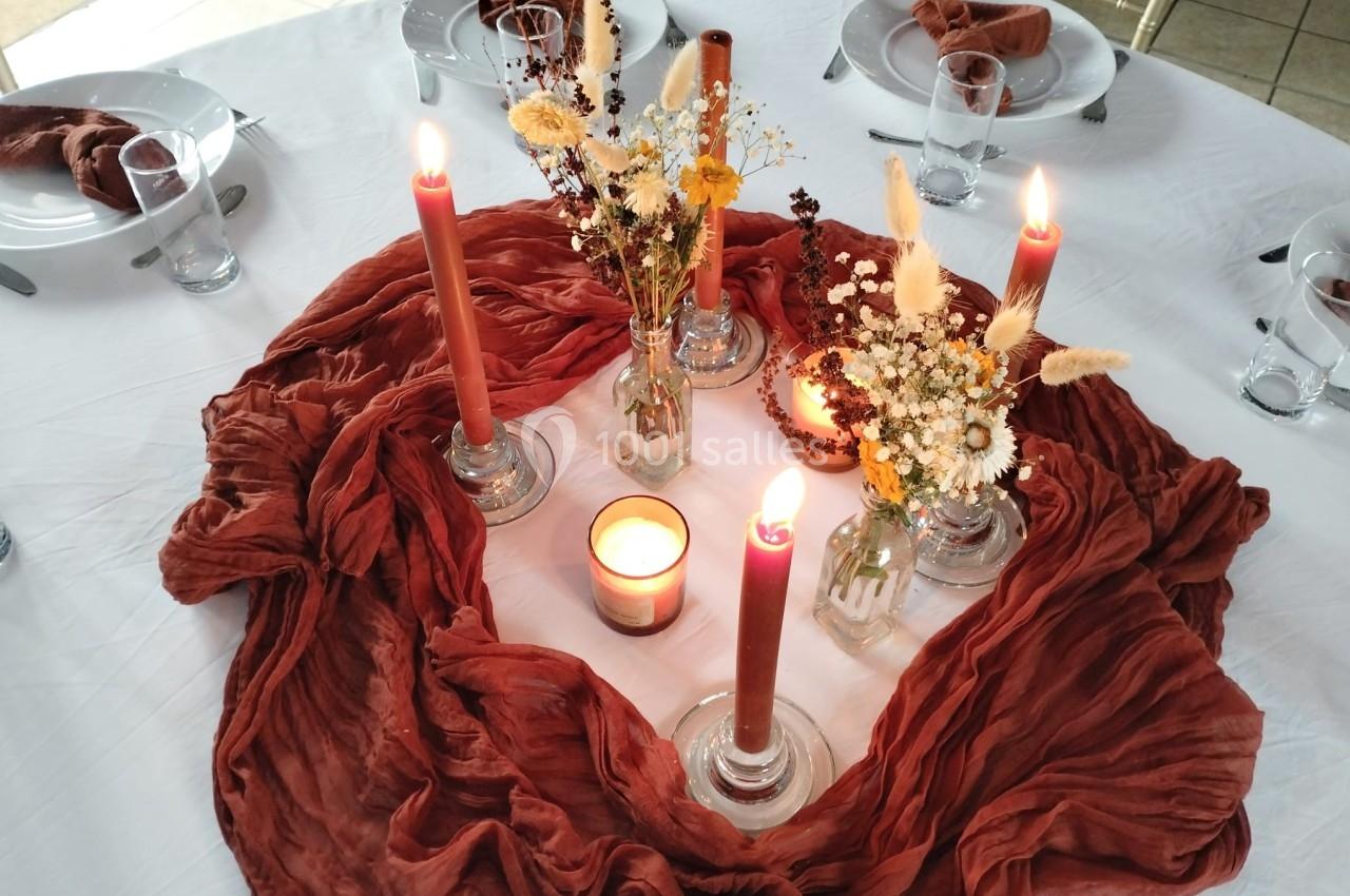 Centre de table avec bougies allumées, fleurs séchées et tissu rouge sur une nappe blanche.