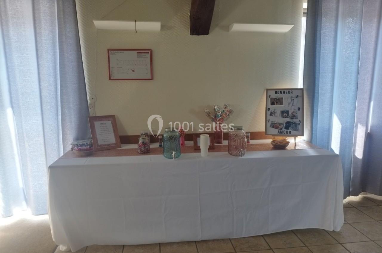 Table décorée avec nappes blanches, bonbonnières, cadre photo et pancartes, placée devant un mur clair avec rideaux bleus.