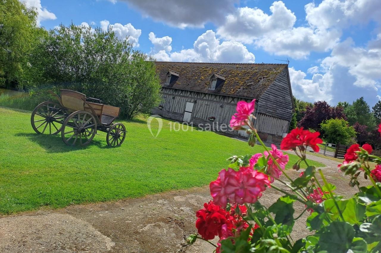 Charrette ancienne en bois sur une pelouse devant une grange à colombages, avec des fleurs rouges au premier plan.