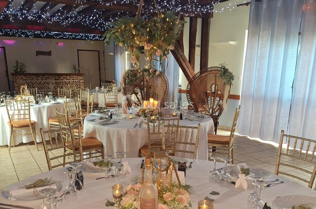 Salle de réception décorée pour un mariage avec tables rondes, chaises dorées, bougies et fleurs.
