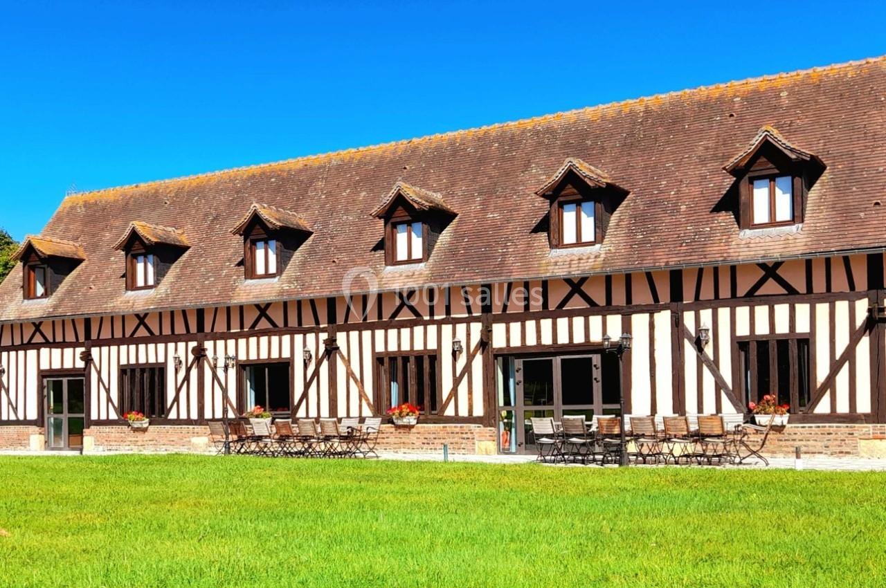Bâtiment à colombages avec toit en tuiles, fenêtres mansardées et terrasse aménagée, entouré de pelouse verte.