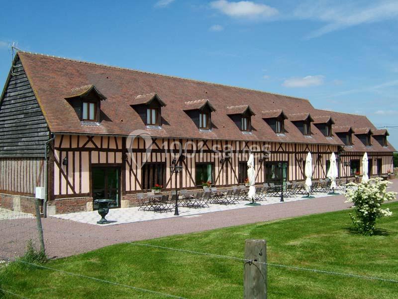 Bâtiment à colombages avec toit en tuiles, fenêtres mansardées et terrasse aménagée, entouré de pelouse et ciel dégagé.