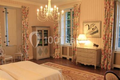 Chambre lumineuse avec un lit double, des rideaux fleuris, un lustre, des meubles classiques et des fenêtres donnant sur la…
